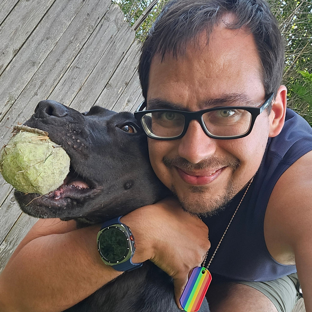 Michael wearing black-rimmed glasses and a navy sleeveless shirt hugs a black dog holding a green ball in its mouth, with a wooden deck and greenery in the background.