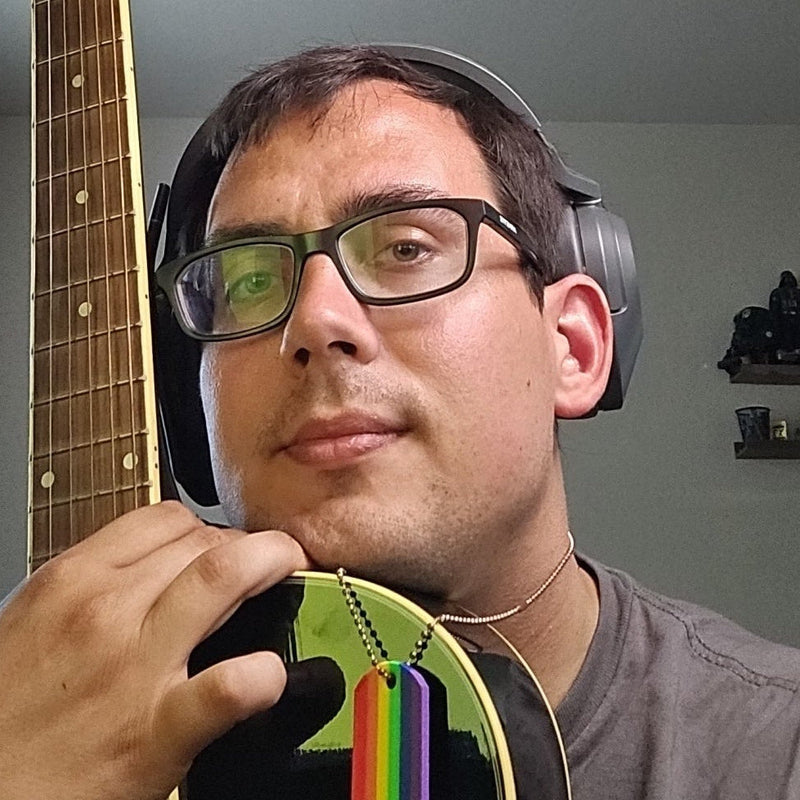 A man wearing glasses and over-ear headphones holds a guitar in a room. The guitar has a black body with a green headstock and a rainbow-colored dog tag pendant.