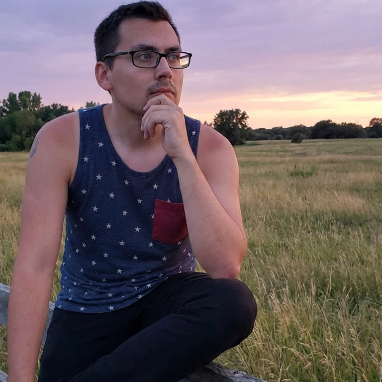 A man wearing glasses, a navy blue sleeveless tank top with white polka dots and a red pocket, and black pants sits on a wooden fence in a grassy field at sunset.